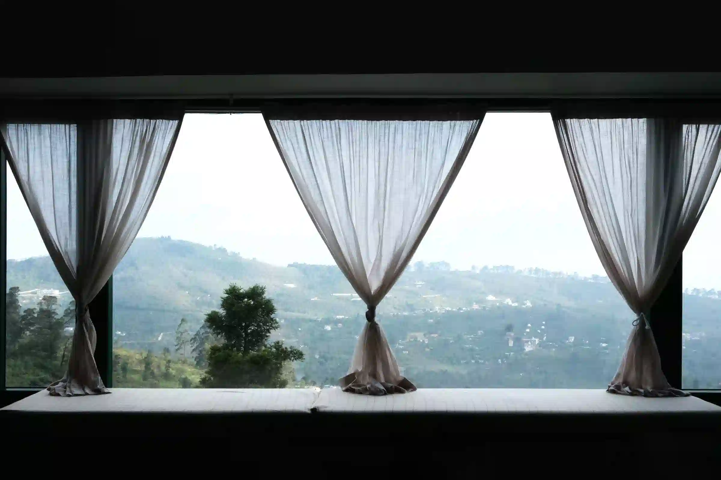 Panoramic mountain view through sheer curtains at Cardamom Pods, featuring a cozy window seat overlooking lush greenery.
