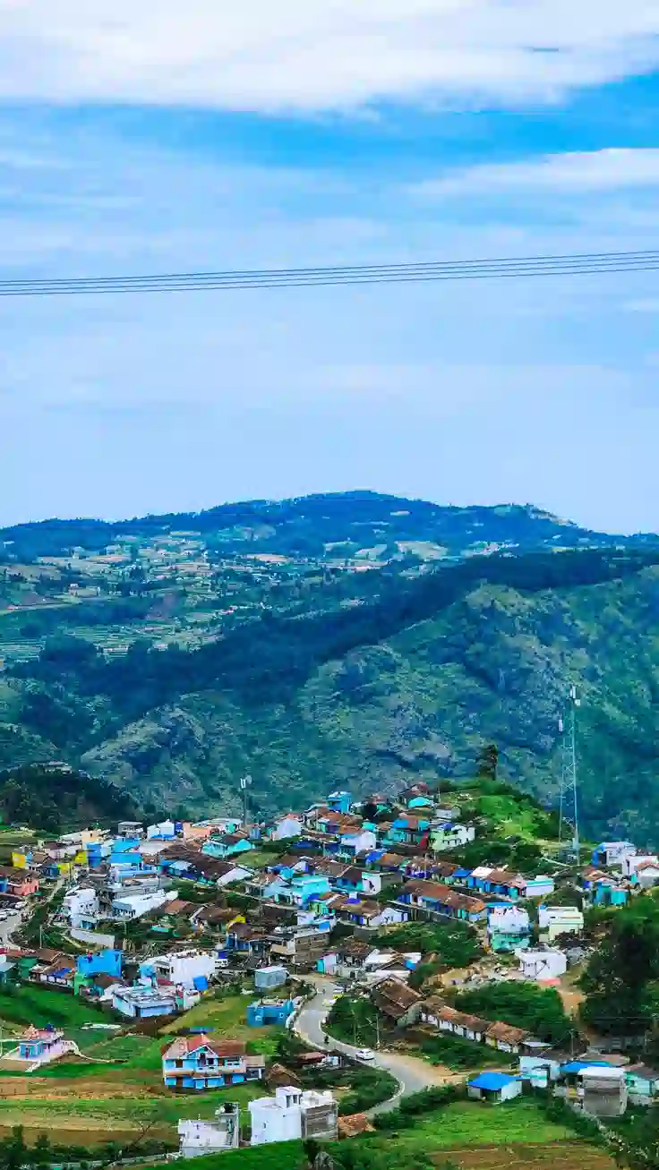 Colorful houses of a hillside village in Kodaikanal nestled among lush green mountains under a bright blue cloudy sky.