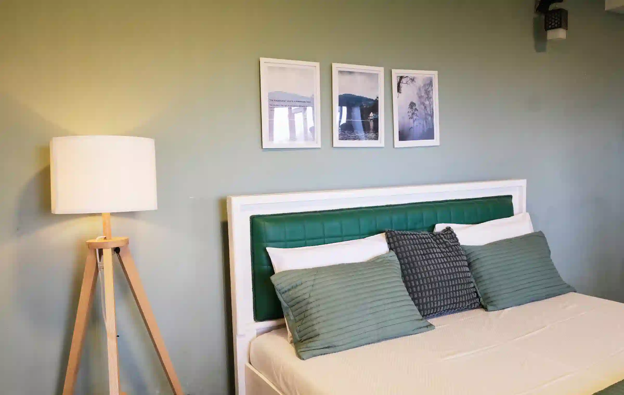 Close-up of double bed at Cardamom Pods showing green textured pillows, warm tripod lighting and scenic framed pictures.